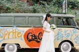A woman in a white dress, possibly a bride, is holding a bouquet of white flowers standing beside a vintage van. The van is painted with graffiti featuring the logos of popular soda brands and some text in another language. The van is parked in an outdoor setting with green foliage visible in the background.