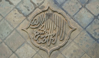 An intricately designed piece of calligraphy is set within a decorative arabesque border, placed on a textured, tiled surface. The calligraphy appears to be carved into the tiles, creating a three-dimensional effect.
