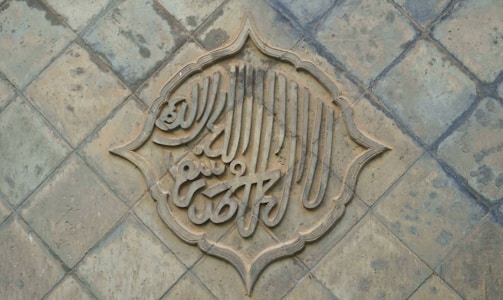 An intricately designed piece of calligraphy is set within a decorative arabesque border, placed on a textured, tiled surface. The calligraphy appears to be carved into the tiles, creating a three-dimensional effect.