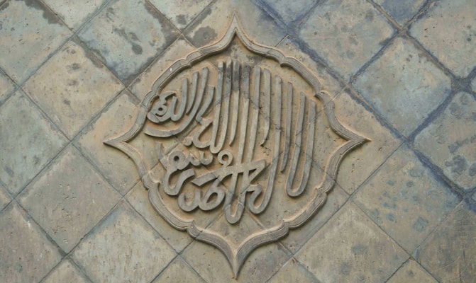 An intricately designed piece of calligraphy is set within a decorative arabesque border, placed on a textured, tiled surface. The calligraphy appears to be carved into the tiles, creating a three-dimensional effect.