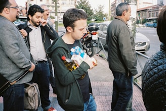 A group of people stand close to the edge of a street. One person is holding several grocery items including a bottle of oil and a carton. The surrounding environment includes buildings, a parked motorcycle, and a car in the background. The atmosphere appears to be casual with people dressed in everyday clothing.