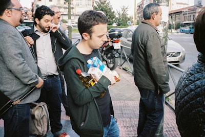 A group of people stand close to the edge of a street. One person is holding several grocery items including a bottle of oil and a carton. The surrounding environment includes buildings, a parked motorcycle, and a car in the background. The atmosphere appears to be casual with people dressed in everyday clothing.
