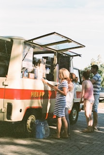 An outdoor setting with a vintage-style ice cream truck parked on a paved surface. The truck has a red stripe and an open service window displaying various ice cream cones and products. Several people, including a woman in a striped dress and others wearing casual summer clothing, are gathered around the truck, possibly ordering or waiting for ice cream.