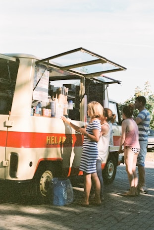 An outdoor setting with a vintage-style ice cream truck parked on a paved surface. The truck has a red stripe and an open service window displaying various ice cream cones and products. Several people, including a woman in a striped dress and others wearing casual summer clothing, are gathered around the truck, possibly ordering or waiting for ice cream.