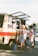 An outdoor setting with a vintage-style ice cream truck parked on a paved surface. The truck has a red stripe and an open service window displaying various ice cream cones and products. Several people, including a woman in a striped dress and others wearing casual summer clothing, are gathered around the truck, possibly ordering or waiting for ice cream.