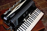A detailed view of a black accordion placed on a wooden surface. The accordion features intricate design elements including multiple rows of buttons and black and white keys. The background includes a patterned carpet with earthy tones, providing a cozy and classic feel.