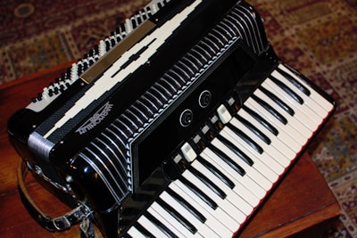 A detailed view of a black accordion placed on a wooden surface. The accordion features intricate design elements including multiple rows of buttons and black and white keys. The background includes a patterned carpet with earthy tones, providing a cozy and classic feel.