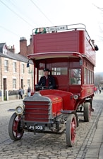 A vintage red double-decker bus on a cobblestone street. The bus has a sign labeled 'Circular' on top and is driven by a person in period clothing. Several passengers are visible, with some seated in the upper deck. The surrounding area features brick buildings and a few pedestrians.