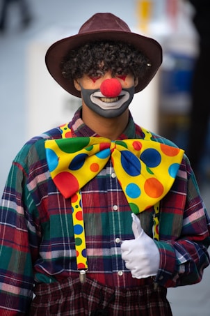 A person dressed as a clown with curly hair, wearing a large brown hat, a red nose, and a painted face featuring exaggerated makeup and a wide smile. The outfit includes a colorful, oversized bow tie with polka dots, a plaid shirt, white gloves, and suspenders. The individual is giving a thumbs-up gesture.