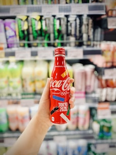 A hand is holding a red Coca-Cola bottle with 'Tokyo 2020' branding, in front of a blurred supermarket shelf filled with various beverage products.