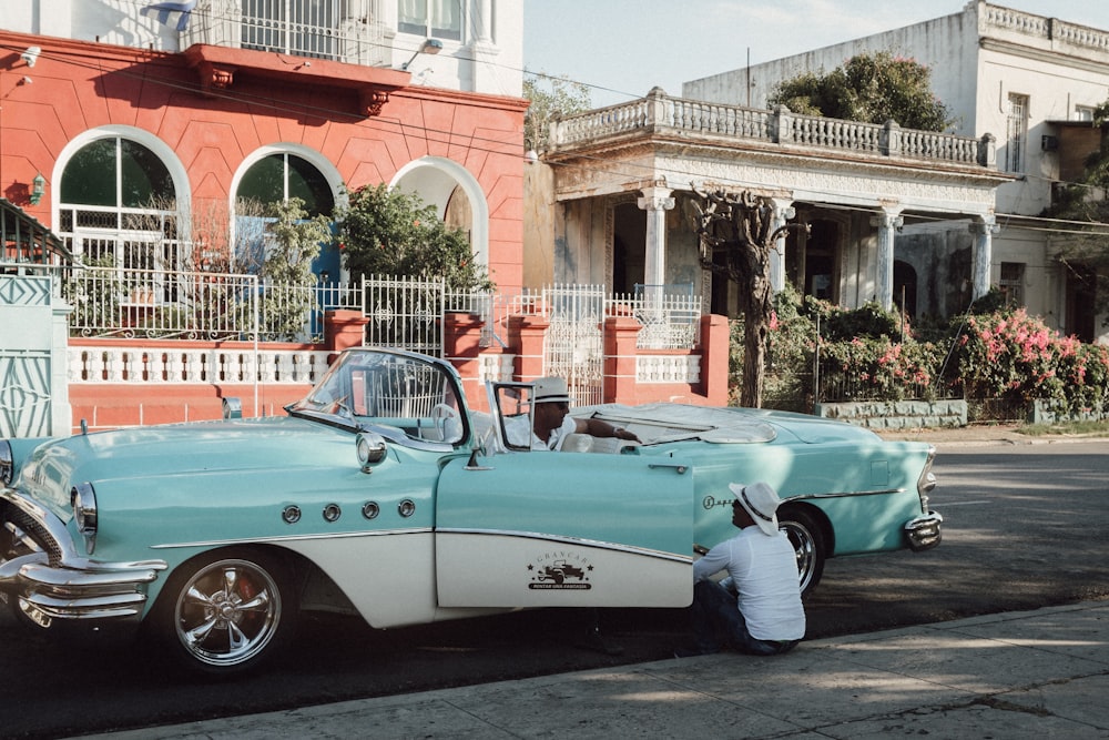 man in white pants and white shirt standing beside teal convertible car during daytime