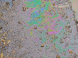 A patch of oil covers a wet asphalt surface, featuring an array of small stones and irregular textures. The oil creates a swirling pattern of iridescent colors including greens, purples, and blues, reflecting light in a rainbow-like effect.