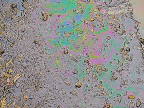 A patch of oil covers a wet asphalt surface, featuring an array of small stones and irregular textures. The oil creates a swirling pattern of iridescent colors including greens, purples, and blues, reflecting light in a rainbow-like effect.