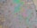 A patch of oil covers a wet asphalt surface, featuring an array of small stones and irregular textures. The oil creates a swirling pattern of iridescent colors including greens, purples, and blues, reflecting light in a rainbow-like effect.
