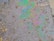 A patch of oil covers a wet asphalt surface, featuring an array of small stones and irregular textures. The oil creates a swirling pattern of iridescent colors including greens, purples, and blues, reflecting light in a rainbow-like effect.