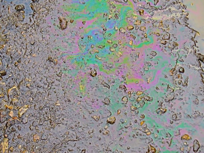A patch of oil covers a wet asphalt surface, featuring an array of small stones and irregular textures. The oil creates a swirling pattern of iridescent colors including greens, purples, and blues, reflecting light in a rainbow-like effect.