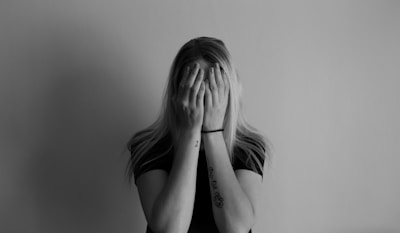 A person with long hair covers their face with both hands against a plain backdrop. They wear a dark shirt and have tattoos on their arms. The orientation and posture suggest feelings of shyness or distress.