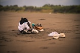 A sandy beach with a collection of items including sandals, water bottles, a backpack, and some clothing piled together. The background features greenery and blurred distant scenery, creating a tranquil atmosphere.