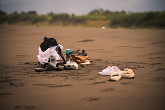 A sandy beach with a collection of items including sandals, water bottles, a backpack, and some clothing piled together. The background features greenery and blurred distant scenery, creating a tranquil atmosphere.