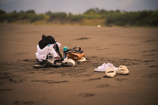 A sandy beach with a collection of items including sandals, water bottles, a backpack, and some clothing piled together. The background features greenery and blurred distant scenery, creating a tranquil atmosphere.