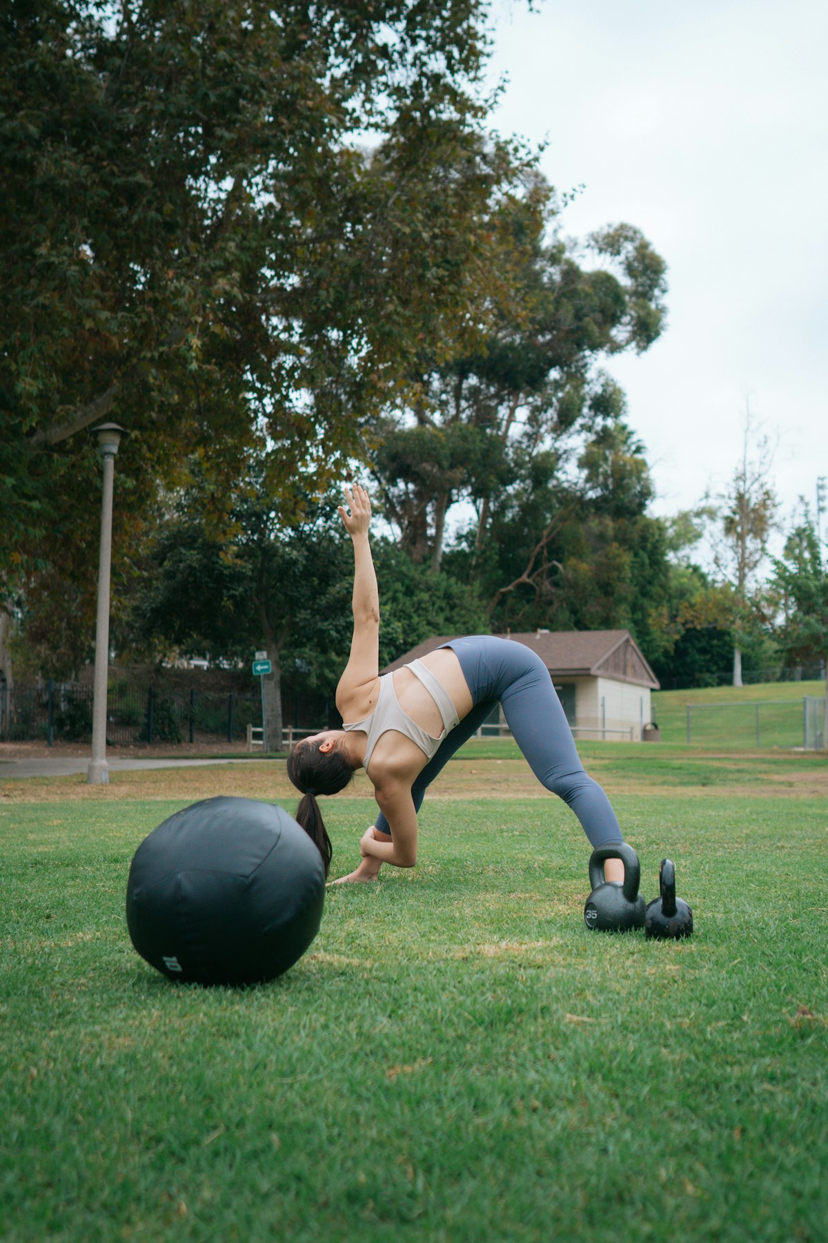 Latihan bodyweight outdoor menggunakan bola stabilitas di taman terbuka menggambarkan gerakan duck walk dan kekuatan kaki ala Primal Movement