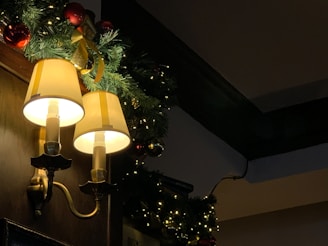 Two illuminated wall-mounted lamps with yellow shades are positioned against a wooden panel. Christmas decorations featuring green pine garland, red baubles, and a gold ribbon are arranged around the lights. Fairy lights add a warm and festive glow to the scene.