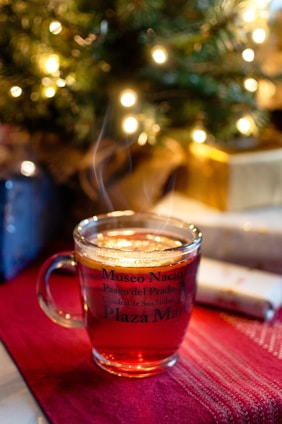 A glass mug filled with a hot beverage, with steam gently rising from the surface. The mug is adorned with text referencing various landmarks, and sits on a deep red cloth. In the background, blurred Christmas lights and what appears to be a decorated tree create a cozy, festive atmosphere, while wrapped gifts are placed nearby.