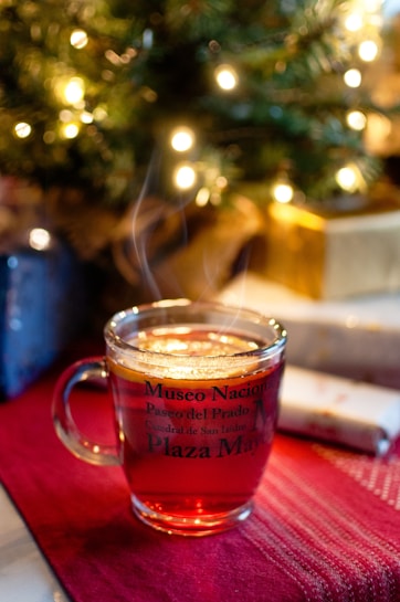 A glass mug filled with a hot beverage, with steam gently rising from the surface. The mug is adorned with text referencing various landmarks, and sits on a deep red cloth. In the background, blurred Christmas lights and what appears to be a decorated tree create a cozy, festive atmosphere, while wrapped gifts are placed nearby.