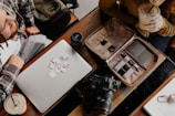 A workspace scene featuring a laptop with multiple memory cards placed on it, a camera, a lens cap, and an organized tech pouch containing cables and pens. Two people are partially visible, one having a drink.