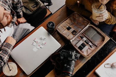 A workspace scene featuring a laptop with multiple memory cards placed on it, a camera, a lens cap, and an organized tech pouch containing cables and pens. Two people are partially visible, one having a drink.