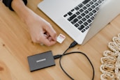 A person is inserting a memory card into a laptop on a wooden table. Next to the laptop, there is an external Samsung hard drive connected via a cable. A woven decorative item is partially visible on the right side.