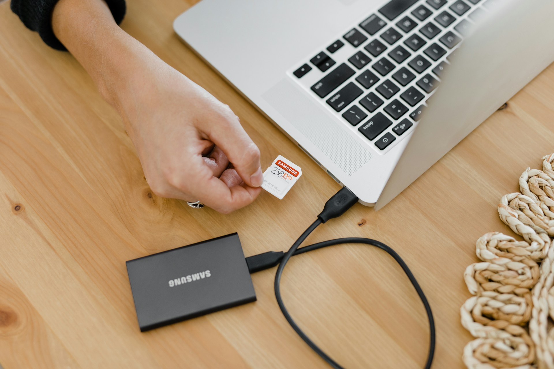 A person is inserting a memory card into a laptop on a wooden table. Next to the laptop, there is an external Samsung hard drive connected via a cable. A woven decorative item is partially visible on the right side.