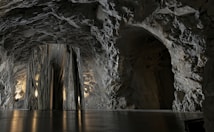 A cave-like environment with rough, rocky walls and ceilings. The lighting is low and warm, emphasizing the texture of the stone. At the center, there is a large, metallic, reflective structure that contrasts with the natural rock surroundings.