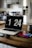 A modern kitchen with a wooden countertop has a MacBook displaying the time 1:24 PM. Beside the laptop, there is a smartphone, wireless earbuds, and a tablet. In the background are stainless steel appliances including a refrigerator and a stove.