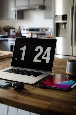 A modern kitchen with a wooden countertop has a MacBook displaying the time 1:24 PM. Beside the laptop, there is a smartphone, wireless earbuds, and a tablet. In the background are stainless steel appliances including a refrigerator and a stove.