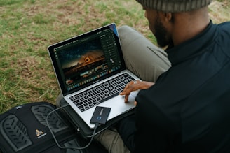 A person is sitting outdoors on grass, using a laptop to edit a photograph. The laptop displays an image of a night scene with light trails. A small external hard drive is connected to the laptop, and a black backpack is nearby.