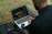 A person is sitting outdoors on grass, using a laptop to edit a photograph. The laptop displays an image of a night scene with light trails. A small external hard drive is connected to the laptop, and a black backpack is nearby.