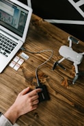 A wooden table displays a laptop with an open screen showing an application, several memory cards, an external hard drive being accessed by a person's hand, and a small drone.
