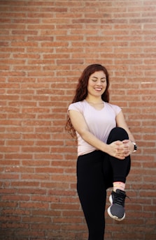 A woman is standing against a brick wall, smiling and holding one knee up with her hands. She is wearing a light pink top and black leggings, along with black sneakers. Her long, wavy hair is loose, and she appears to be in a relaxed pose as if stretching.