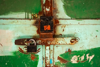A rusty metal latch and lock are affixed to a weathered, painted green door, showing signs of decay and wear. The patches of rust and faded paint create an aged, rustic appearance, with small areas of chipped paint revealing the metal underneath.