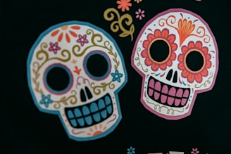 Two colorful sugar skulls decorated with intricate floral and swirl designs are prominently featured against a dark background. The skulls have patterns of flowers, swirls, and decorative elements characterized by bright colors such as pink, orange, blue, and yellow.