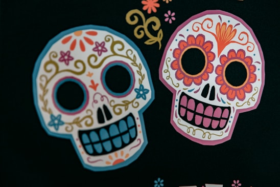 Two colorful sugar skulls decorated with intricate floral and swirl designs are prominently featured against a dark background. The skulls have patterns of flowers, swirls, and decorative elements characterized by bright colors such as pink, orange, blue, and yellow.