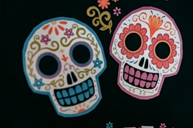 Two colorful sugar skulls decorated with intricate floral and swirl designs are prominently featured against a dark background. The skulls have patterns of flowers, swirls, and decorative elements characterized by bright colors such as pink, orange, blue, and yellow.