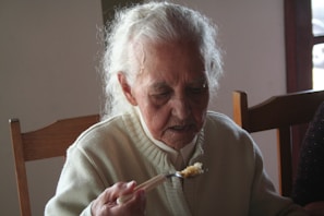 An elderly person with grey hair is seated at a wooden table, wearing a cream-colored sweater and holding a fork with food on it. The setting appears to be indoors with natural light coming from a window.
