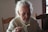 An elderly person with grey hair is seated at a wooden table, wearing a cream-colored sweater and holding a fork with food on it. The setting appears to be indoors with natural light coming from a window.