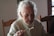 An elderly person with grey hair is seated at a wooden table, wearing a cream-colored sweater and holding a fork with food on it. The setting appears to be indoors with natural light coming from a window.