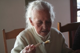 An elderly person with grey hair is seated at a wooden table, wearing a cream-colored sweater and holding a fork with food on it. The setting appears to be indoors with natural light coming from a window.