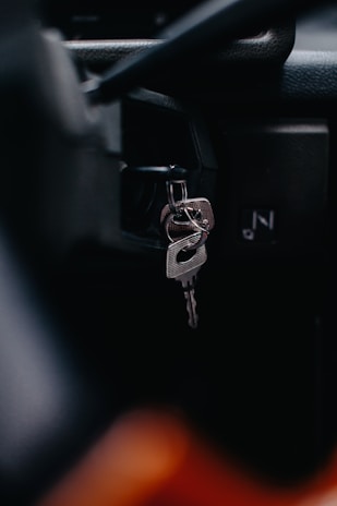 A close-up view of a car's ignition switch with keys inserted. The focus is on the metal keychain and key, with a blurred background that suggests a vehicle interior.