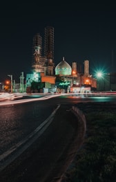 A nighttime scene features a mosque under construction, with scaffolding around its minarets. The building is illuminated with green and red lights, creating an atmospheric effect. In the foreground, light trails from passing vehicles blur along a curved road, enhancing the sense of movement.