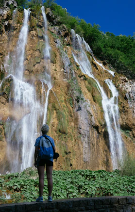 man in blue jacket and black backpack standing on green grass field near waterfalls during daytime
